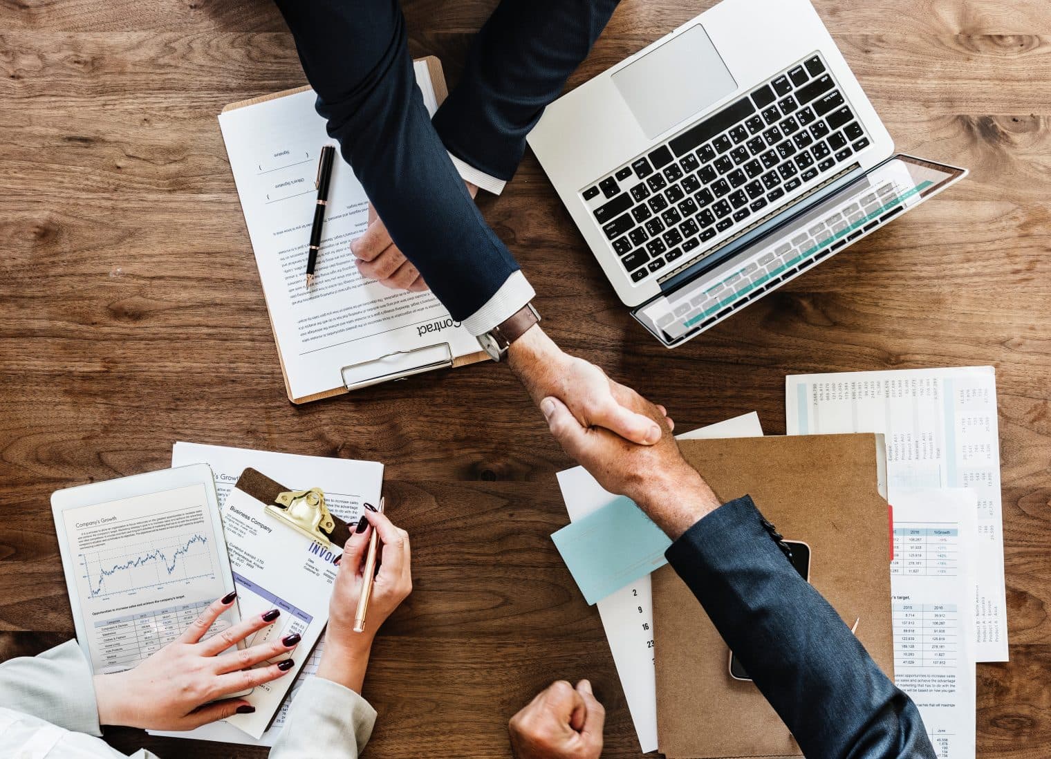 Two men shaking hands in a meeting with papers, and computers in view. Birds eye view.