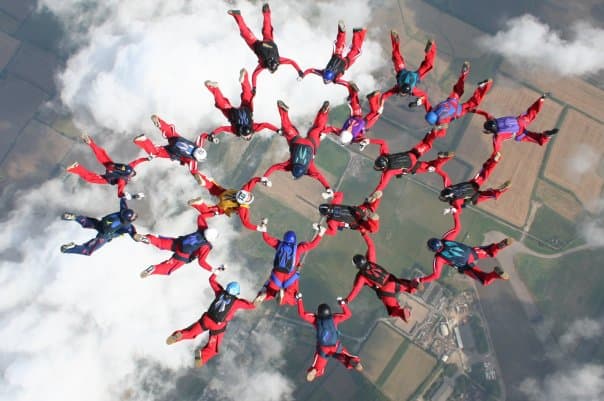 A group of over 20 skydivers wearing red suits and holding hands in a circle formation above the clouds.