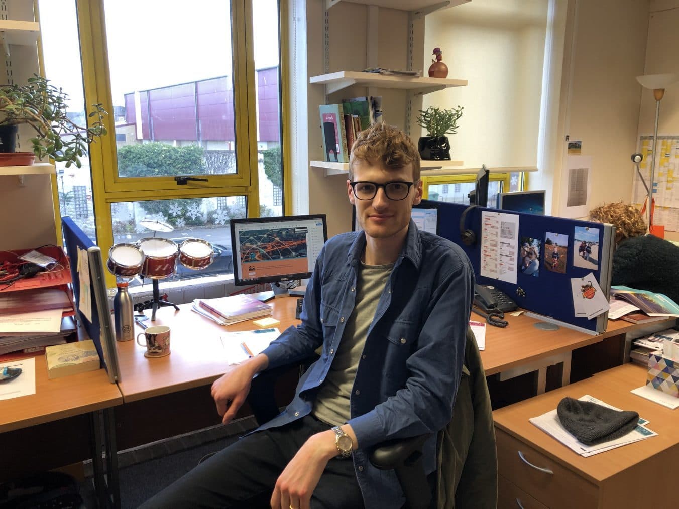 Chris, a man in his late 20s with curly ginger hair, at his desk and smiling at the camera.