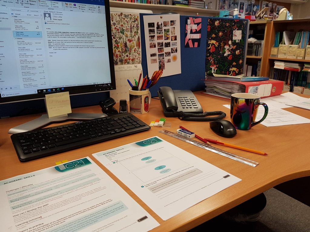 Alice's desk with papers, pens, folders, and a cup of tea. Emails are open on her computer screen.