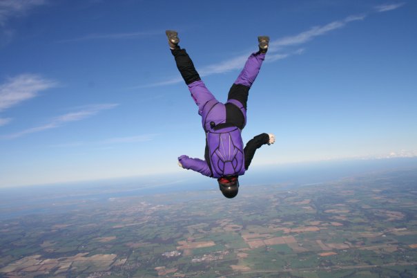 A woman in a purple suit in free-fall head first over a scenic countryside view of Kent, England. 