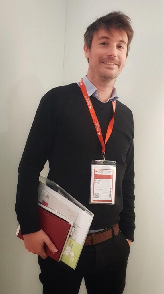 Matt, a tall, smiling man, wearing a conference lanyard, and holding papers and folders ready to go to a meeting. 