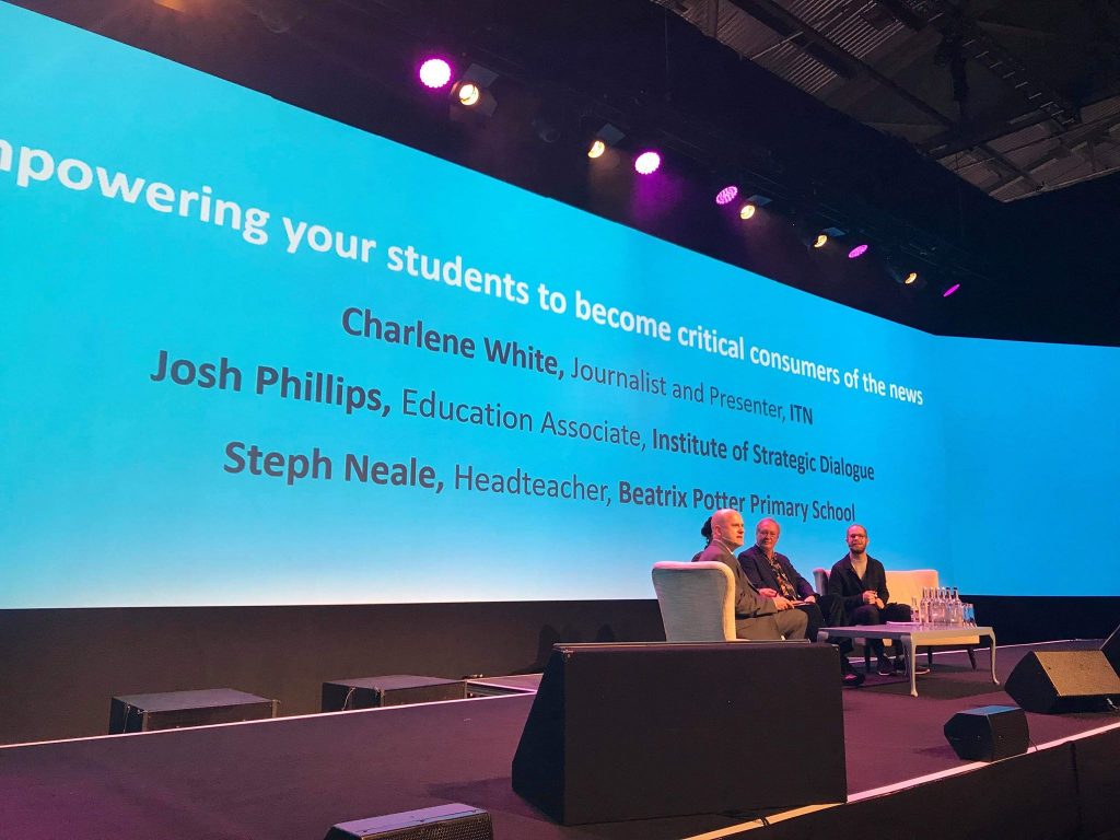 A collection of people sat on sofas in the middle of a stage with a big blue screen behind them. The talk is about Empowering your students to become critical consumers of the news.
