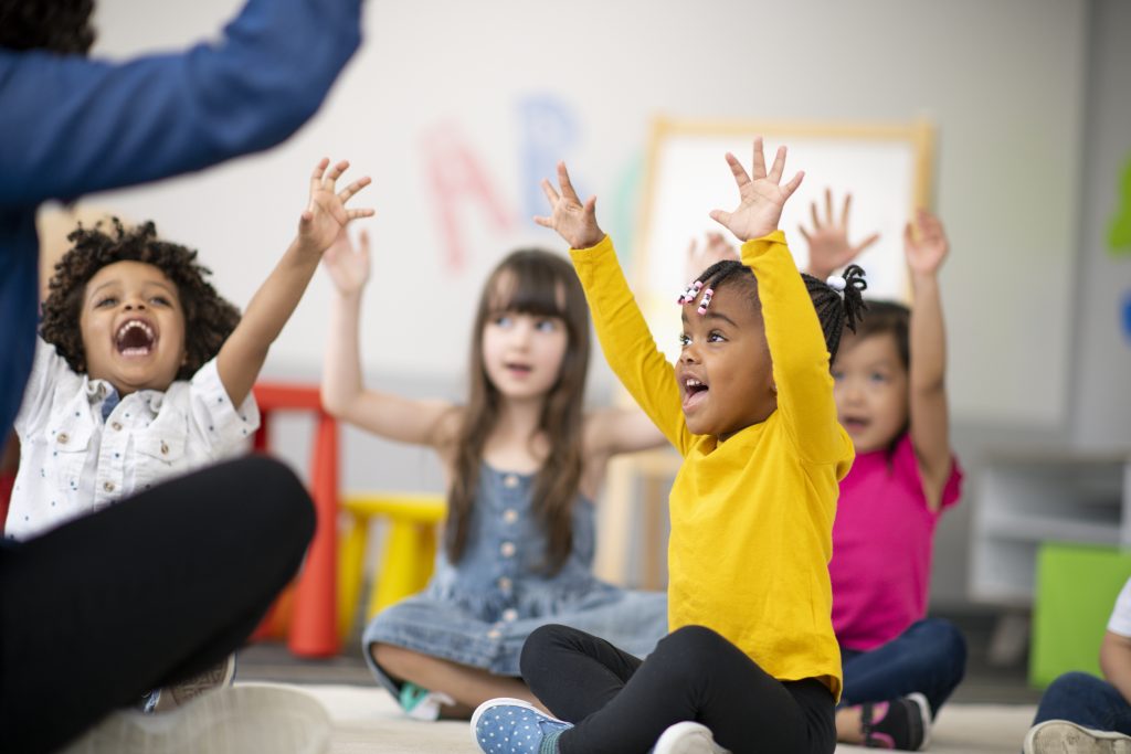 A multi-ethnic group of preschool students is sitting with their legs crossed on the floor in their classroom. The mixed-race female teacher is sitting on the floor facing the children. The happy kids are smiling and following the teacher's instructions. They have their arms raised in the air.