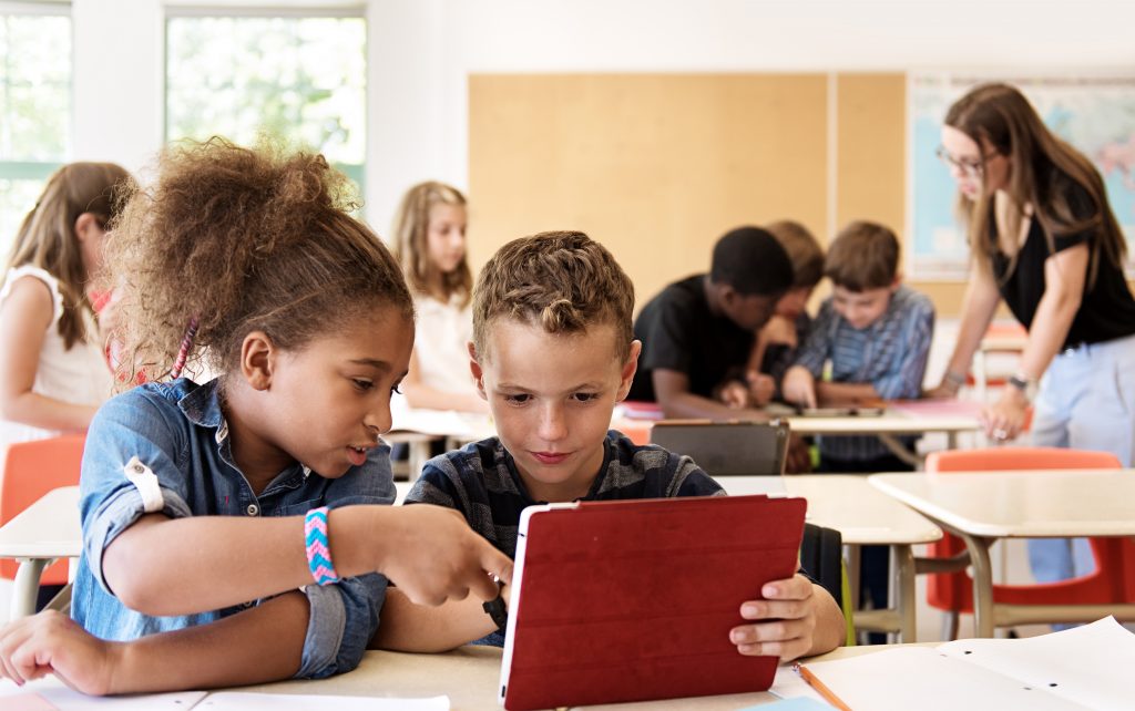 Group of children in a classroom. Focus is on on a young white boy holding a red iPad, and a young black girl is tapping on the screen. 