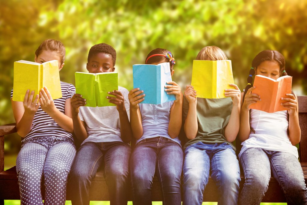A group of children sat on a bench outside, holding up books and reading them. 