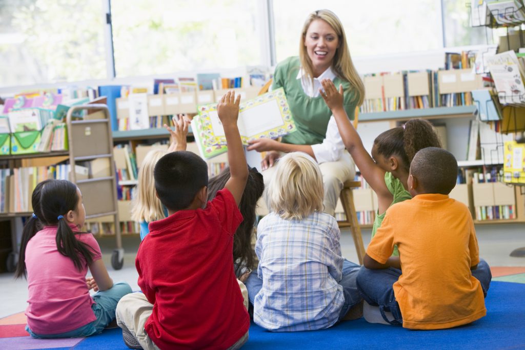 A teacher reading a book to a group of children sat on the floor in a classroom. The students are engaging and putting their hands up. 