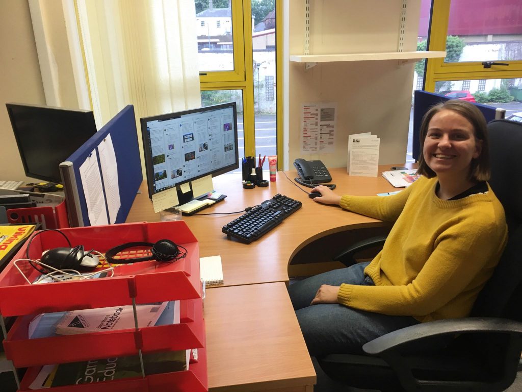 Jaz, a young woman with short brown hair and a mustard yellow jumper, sitting at her desk and smiling at the camera. 